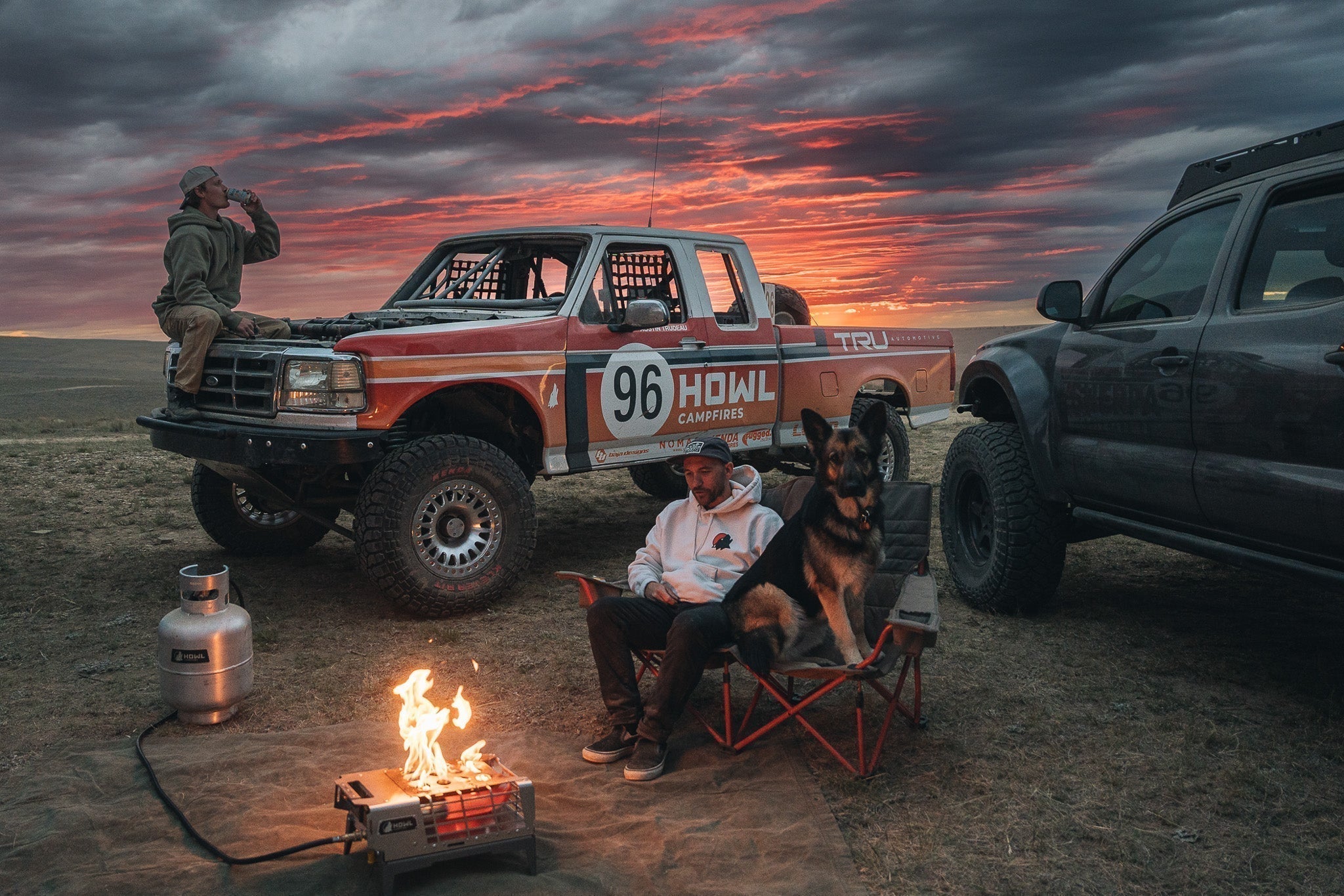 Man and dog sitting beside a HOWL propane campfire in front of an off-road race truck at sunset