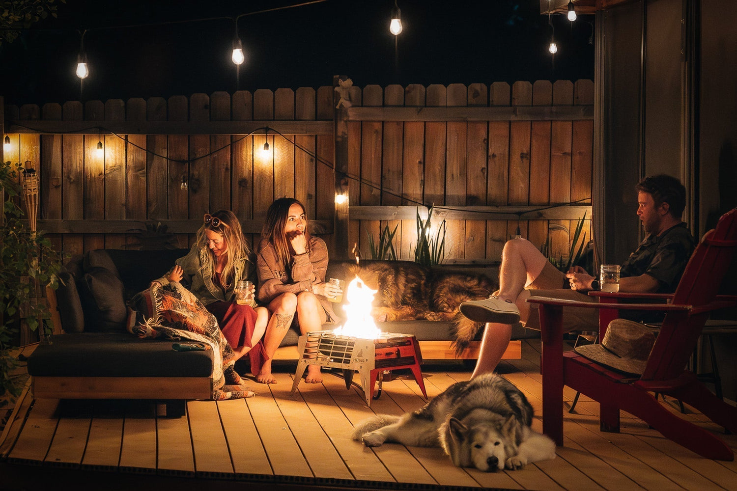 Friends and dogs relaxing around a HOWL propane fire pit on a backyard deck at night.