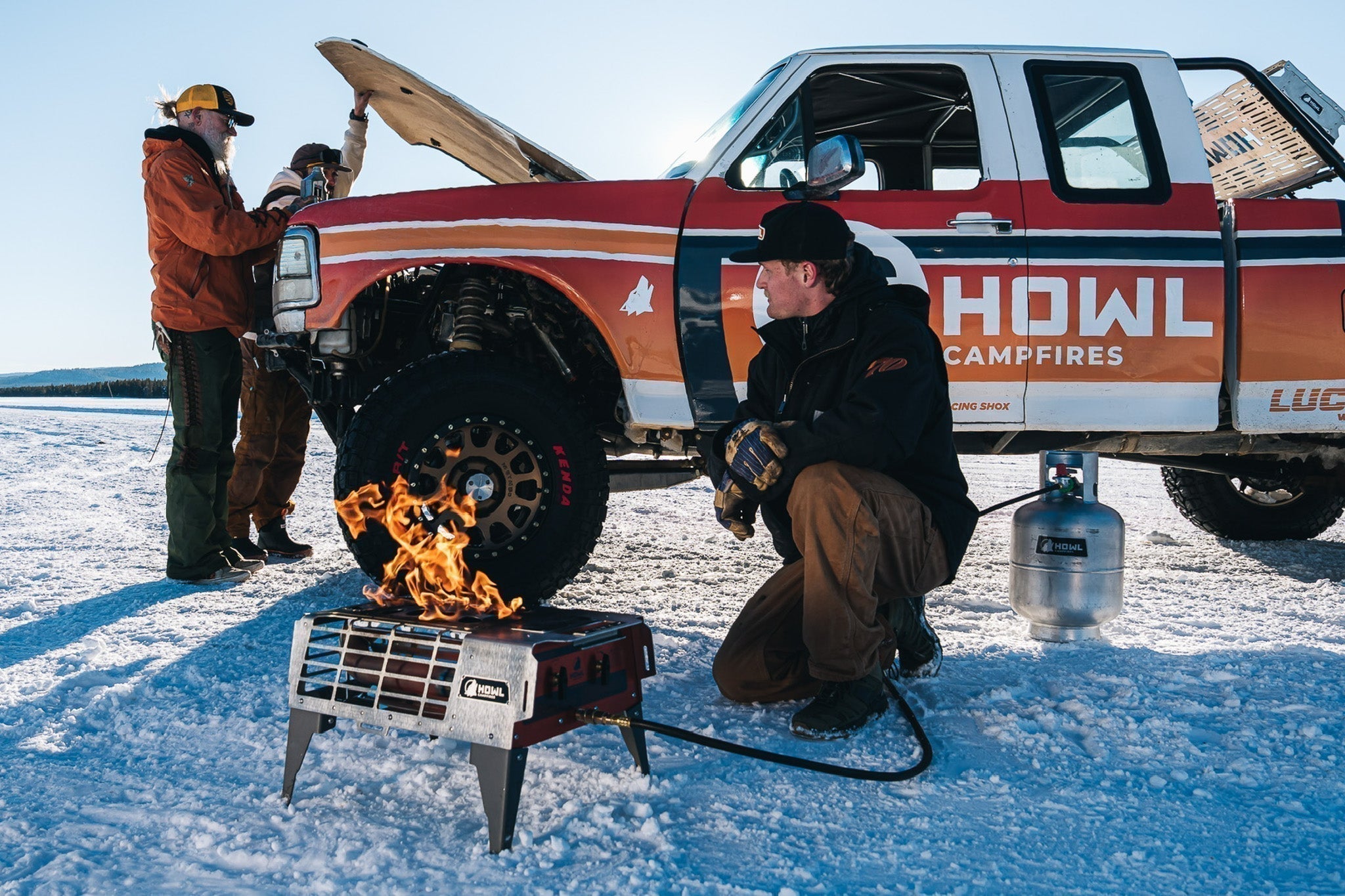 Two campers use a HOWL propane campfire beside a pickup truck in a snowy mountain landscape.