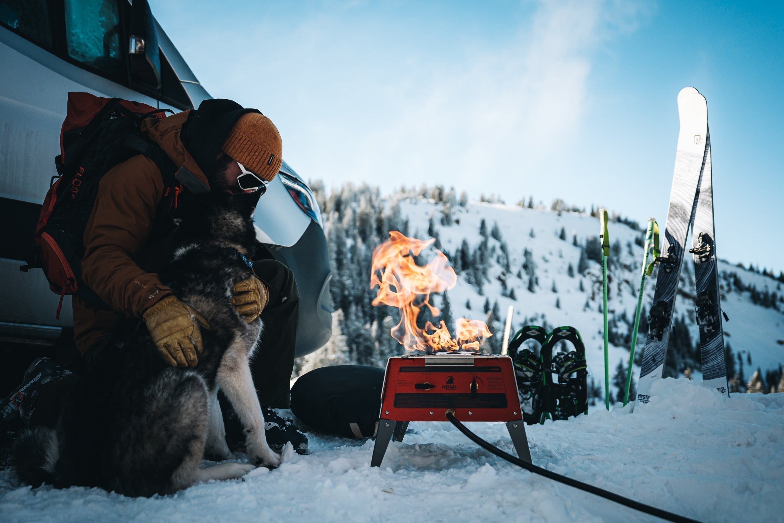 Backcountry skier and dog warming beside a HOWL propane campfire in the snowy mountains.