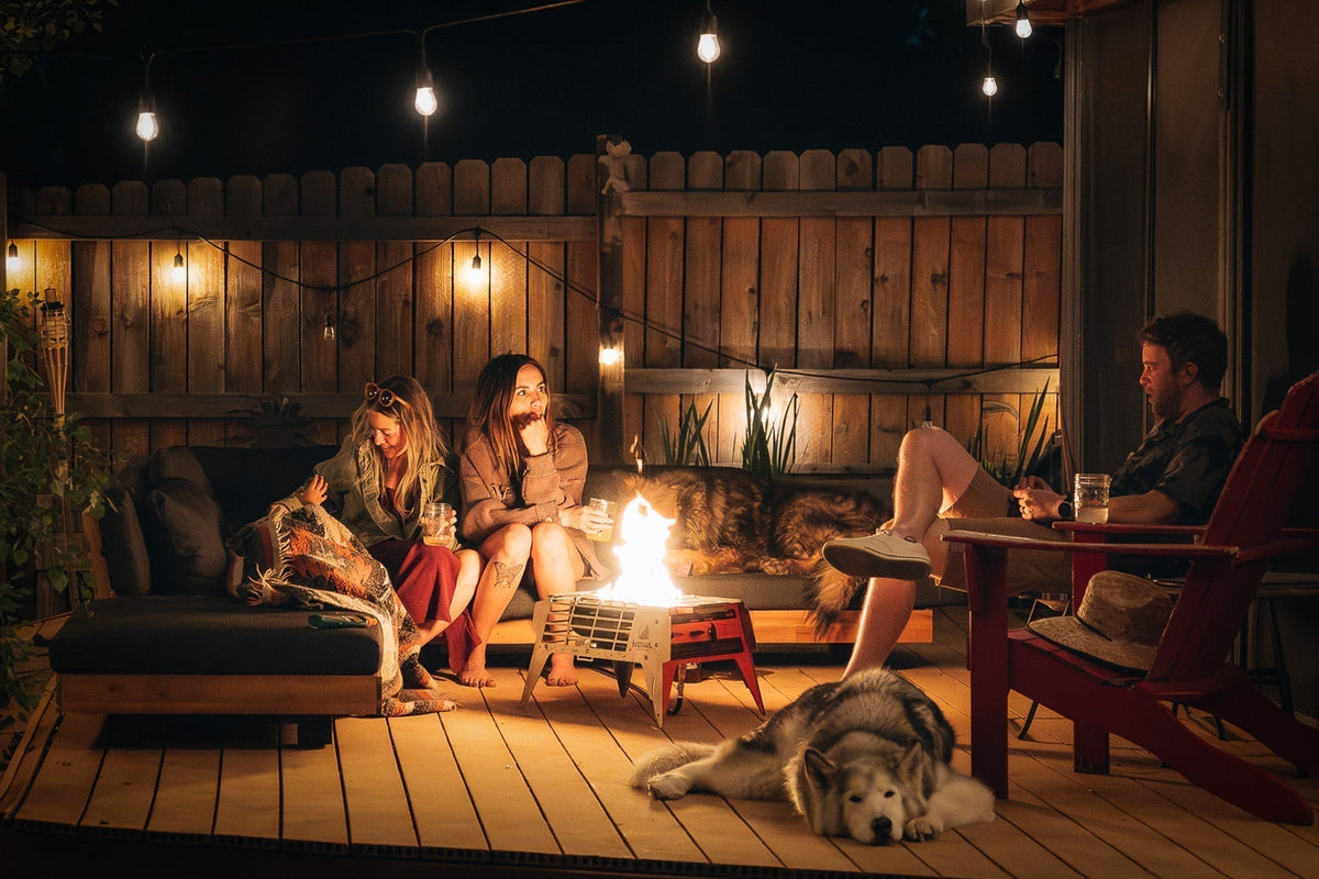 Friends and dogs relaxing around a HOWL propane fire pit on a backyard deck at night.